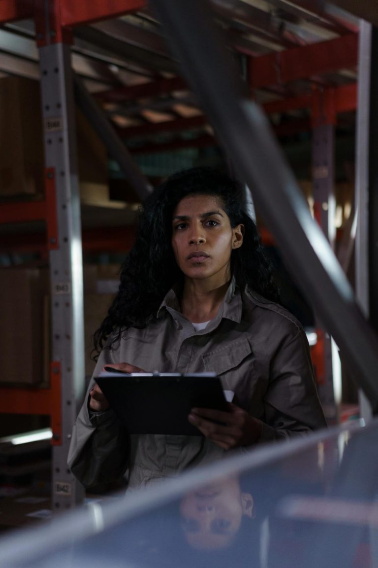 A focused woman holding a clipboard conducting an inventory check in a warehouse aisle.